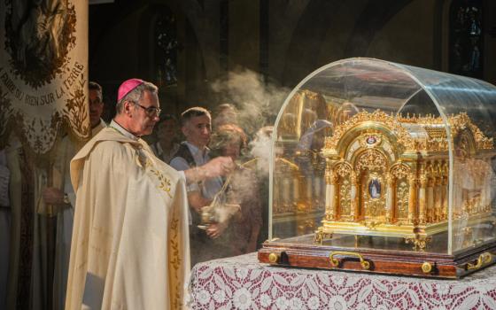 A white man wearing a purple zucchetto and golden vestments swings a thurible towards a gold container