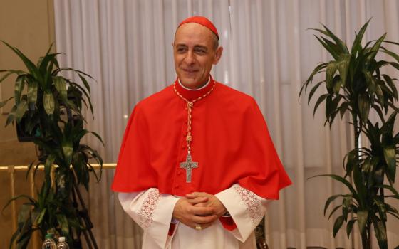 A light-skinned man wears a red cassock and zucchetto and folds his hands in front of him