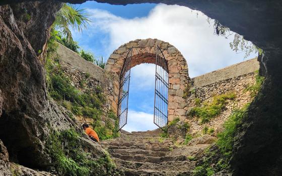 A view from a cenote/cave looks up into the sky through a gated archway in Maní, Yucatan, Mexico (Unsplash/A T Ø M E)