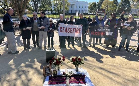 Group stands in front of a fence. White House is visible behind the fence. A person holds a sign that says, "Ceasefire."