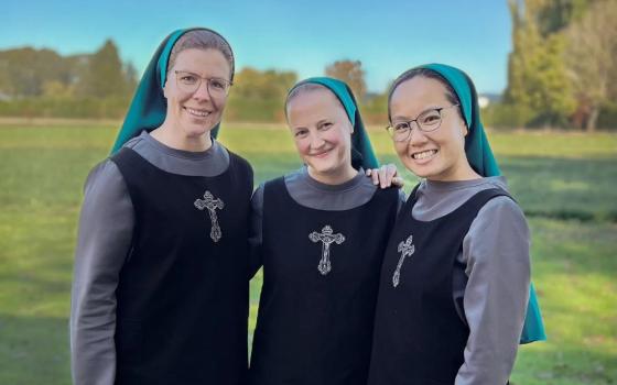 Three women stand together in a field. They are wearing back and gray dress with a cross on the front and teal veils. 