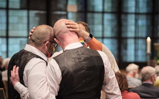 Two men with arms around each pther are seen as a priest puts his hand on nne of their heads in a blessing.