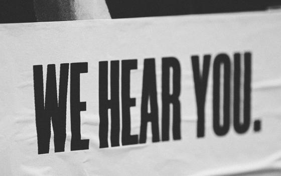 A black-and-white image features a sign with all-caps text that reads: "We hear you." (Unsplash/Jon Tyson)