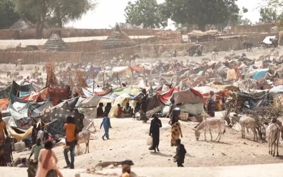 View of many people walking and sitting among tents in dusty area. 