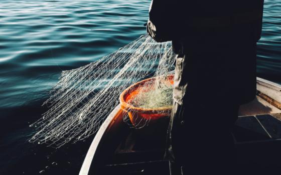 Dark silhouette of a person on a small boat in deep blue waters pulling a white fishing net into an orange bucket.