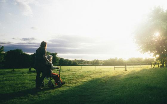 A person pushes an older woman in a wheelchair in a grassy field, with the sun shining upon them, in an outside setting. (Unsplash/Dominik Lange)