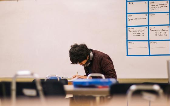 A young man works in a classroom setting. He is seated amid tables and chairs, with a dry erase board behind him. (Unsplash/Jeswin Thomas)