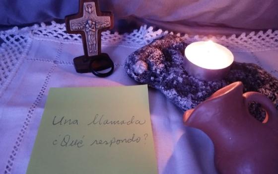 A cross, a clay jug and a candle sit beside a note in Spanish reading, "A call…how shall I respond?" (Flight in V)