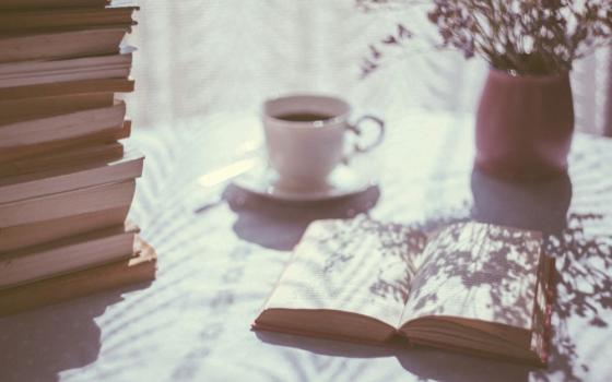 A pile of books, an open book, a cup of coffee or tea, and a plant sit on a table near a window.