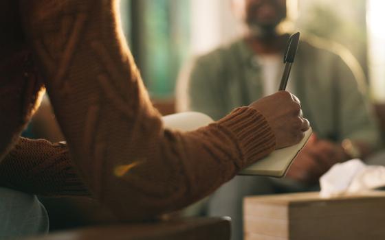 Focus on a therapist's hand holding a pen over notes, with a client in the background (Dreamstime/Yuri  Arcurs)
