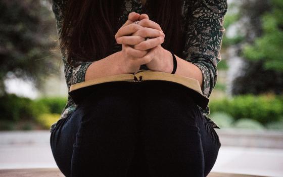 (Unsplash/Olivia Snow) A woman sits with her hands folded on top of what looks to be a Bible on her lap in her outdoor setting. Her face is not visible. (Unsplash/Olivia Snow)