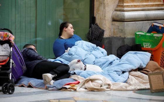 An adolescent boy lying on blanket on the street next to a reclining woman and bags of possessions