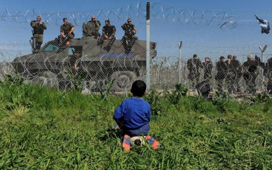 A refugee boy sits on the Greek side of the border near Idomeni as Macedonian police stand guard on their side April 12. See point three below: Pope heads to Greece. (CNS/Alexandros Avramidis)