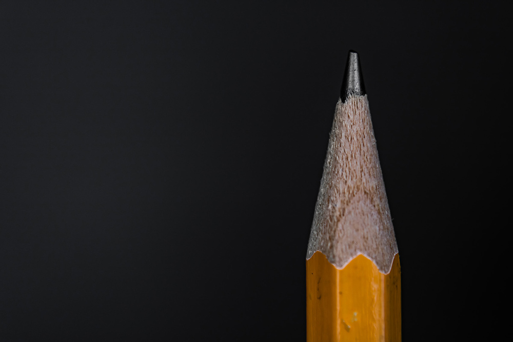 Detalle de la punta de un lápiz y parte de su cuerpo de madera, pintada de amarillo, sobre un rectángulo negro. 